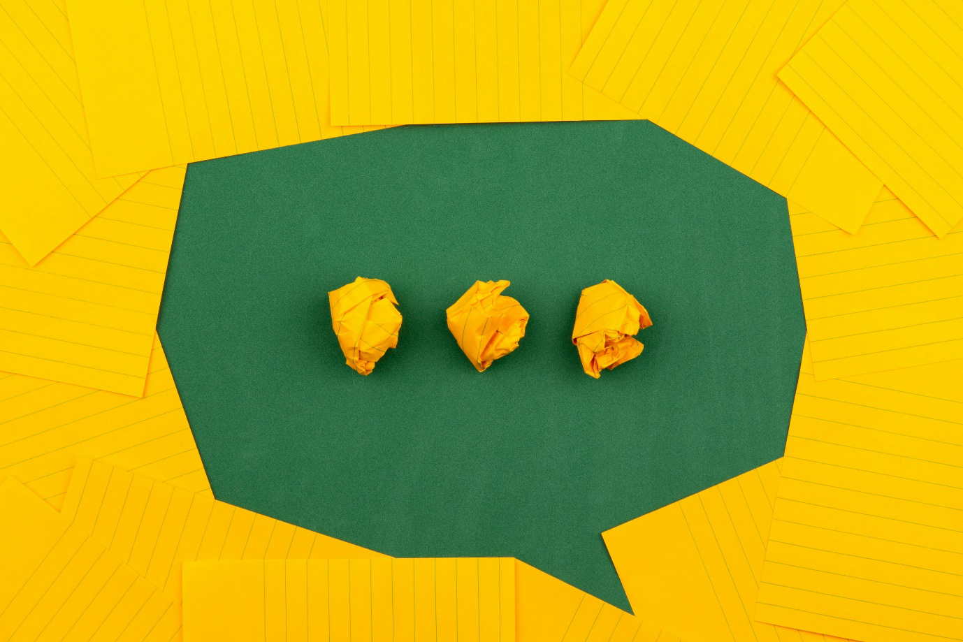 Crumpled yellow papers surrounded by lined sheets on a green background, forming a chat bubble with three dots in it